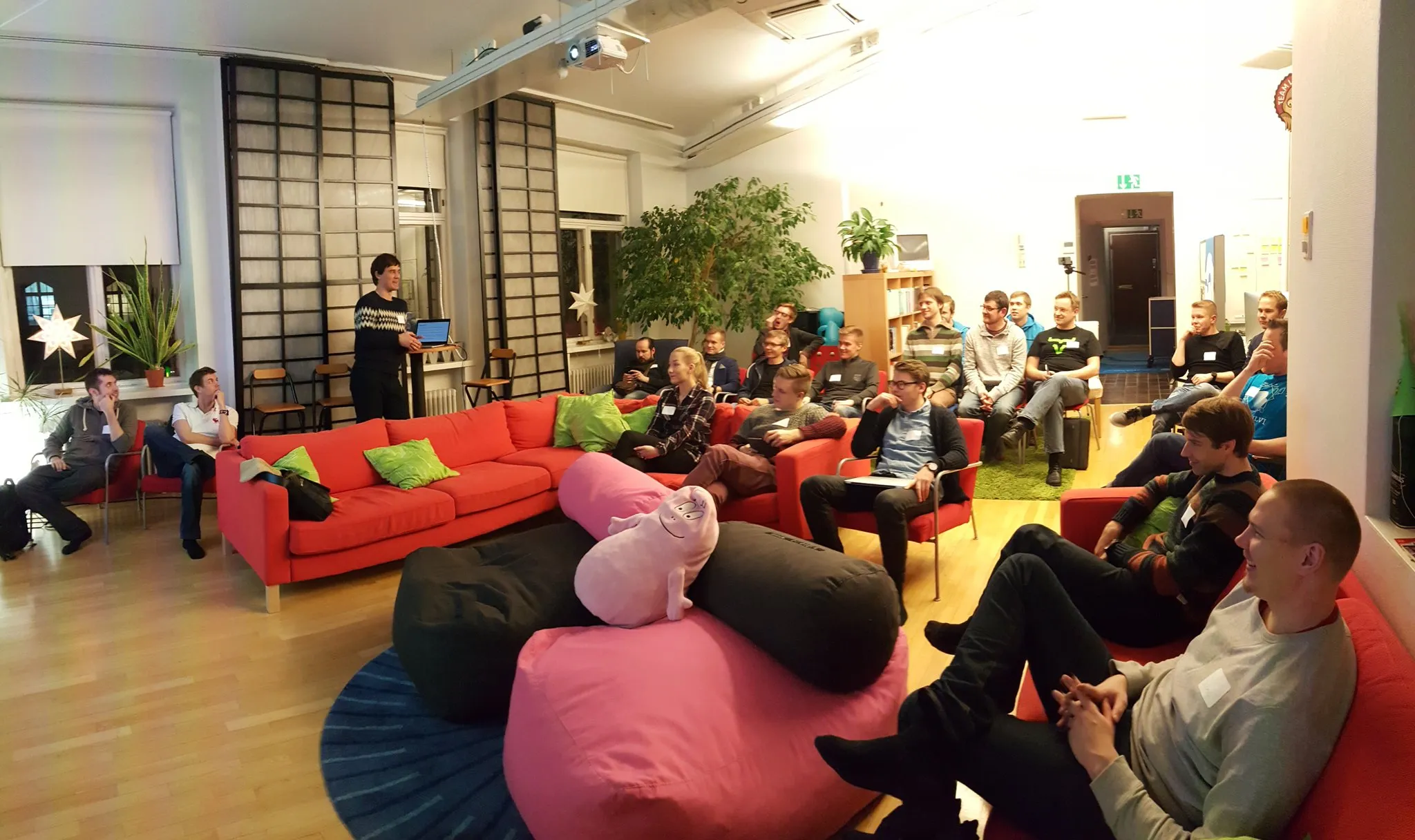 Two dozen people sitting at an office space, some on couches and others on chairs listening to a speaker who's standing to one side with his laptop.