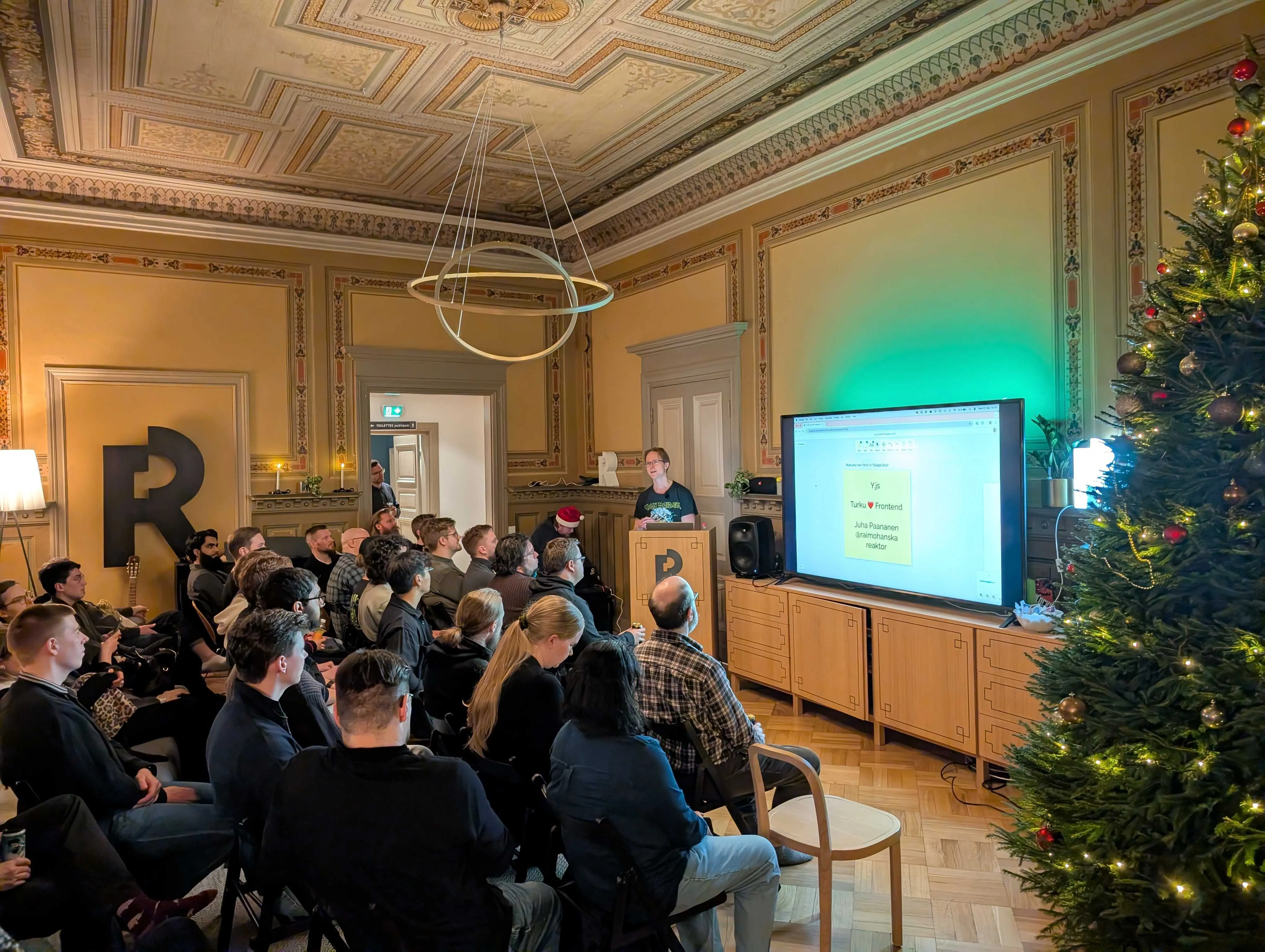 A group of people sitting in a beautifully decorated old room with a Christmas tree and a man speaking next to a large TV